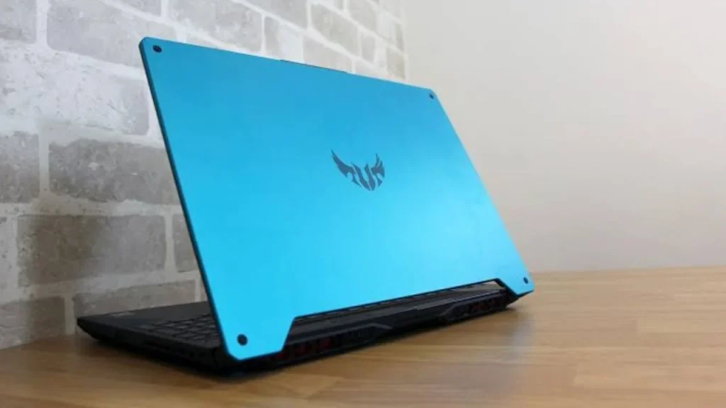 An ASUS TUF Gaming A15 laptop shown from a rear angle, highlighting its vibrant cyan-blue lid and back panel with the TUF logo, sitting on a wooden surface against a light grey brick wall.