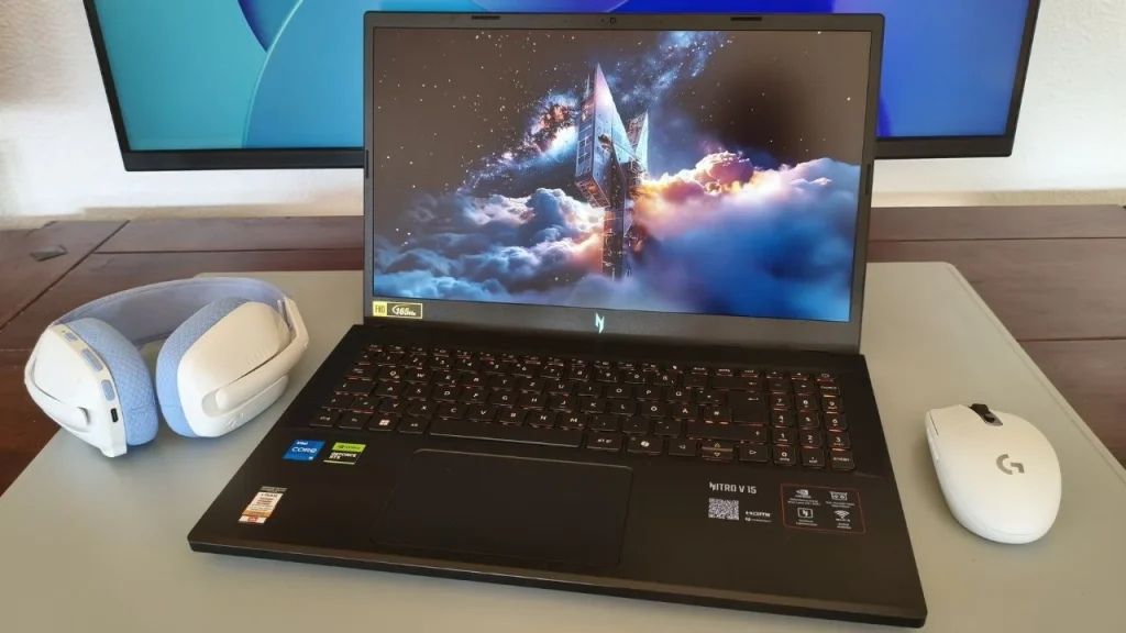 An open Acer Nitro V 15 laptop resting on a desk with a light-colored desk mat, flanked by white-and-blue gaming headphones on the left and a white gaming mouse on the right, with a dual-monitor setup visible in the background.