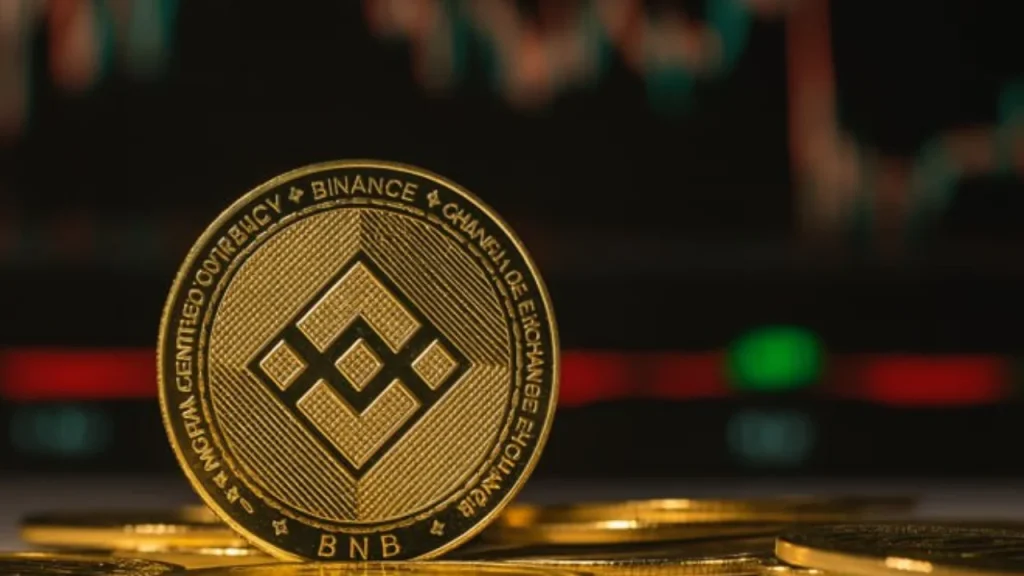 Gold BNB (Binance Coin) coin with the diamond logo standing on a stack of coins, with a blurred red and green trading chart in the background.