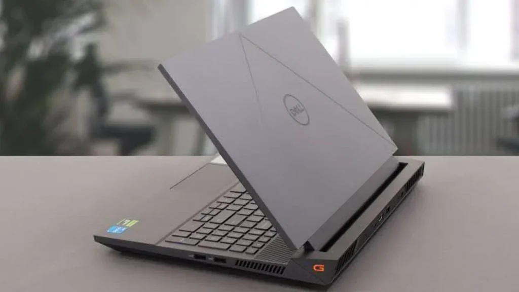 A Dell G15 gaming laptop angled open on a light grey desk, showing its grey lid with faint geometric accent lines and the Dell logo, with a blurred indoor office background.