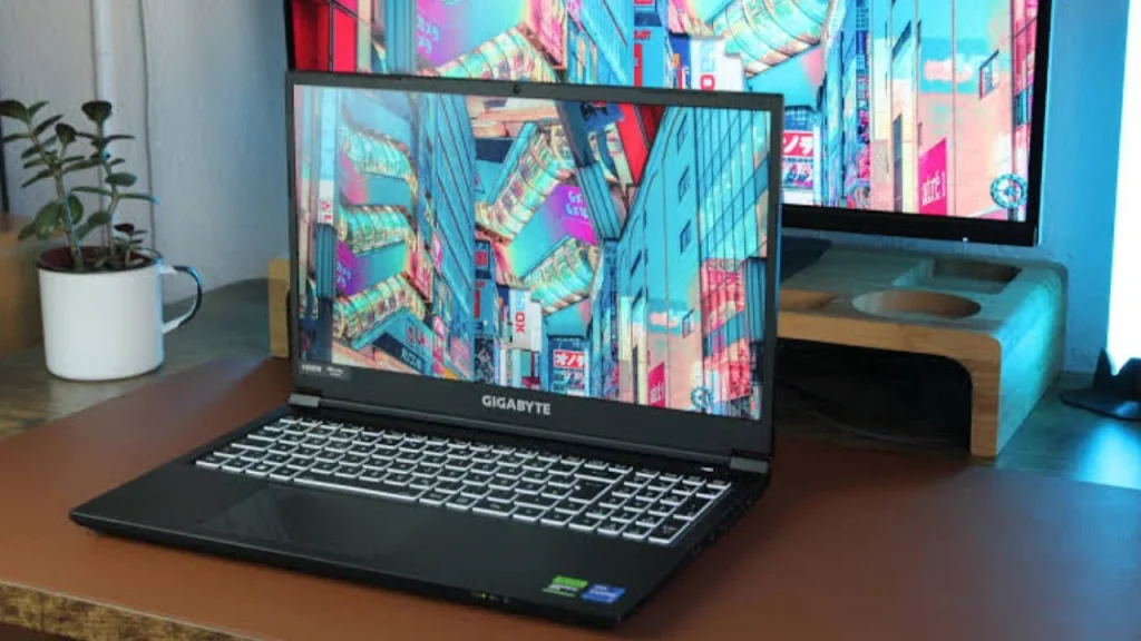 An open Gigabyte G5 gaming laptop resting on a wooden desk, featuring a colorful city street wallpaper on its display, a white mug with a small plant to the left, and a dual-monitor setup in the background.