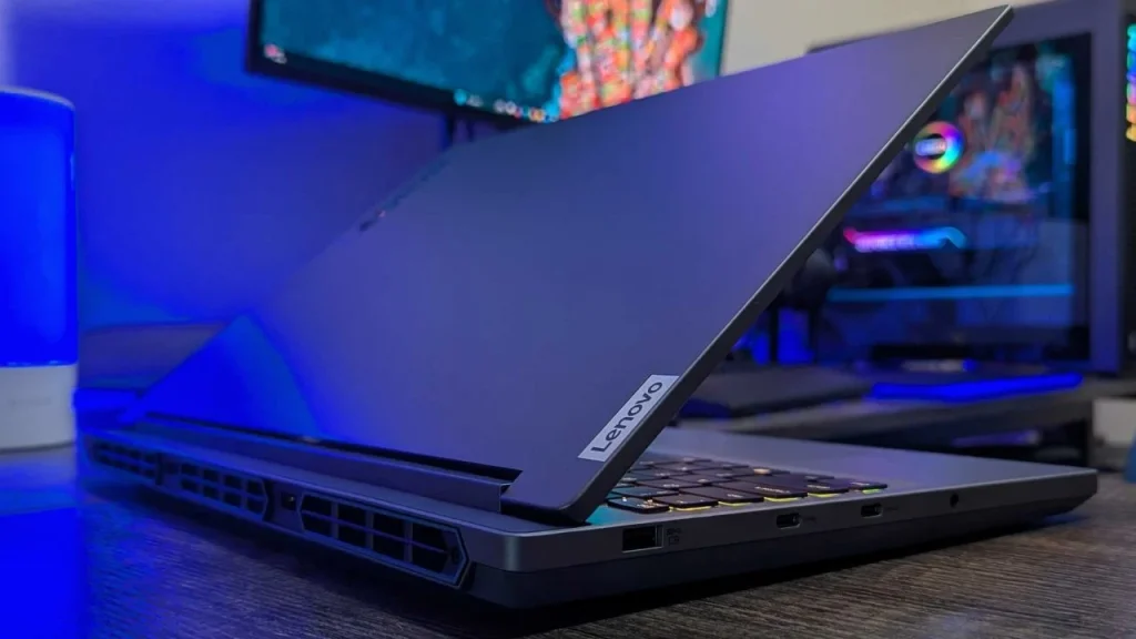 A Lenovo Legion 5i gaming laptop viewed from the rear side, showcasing its dark grey chassis, rear ventilation ports, and the Lenovo logo tag on the hinge, set against a blue-lit gaming setup background.