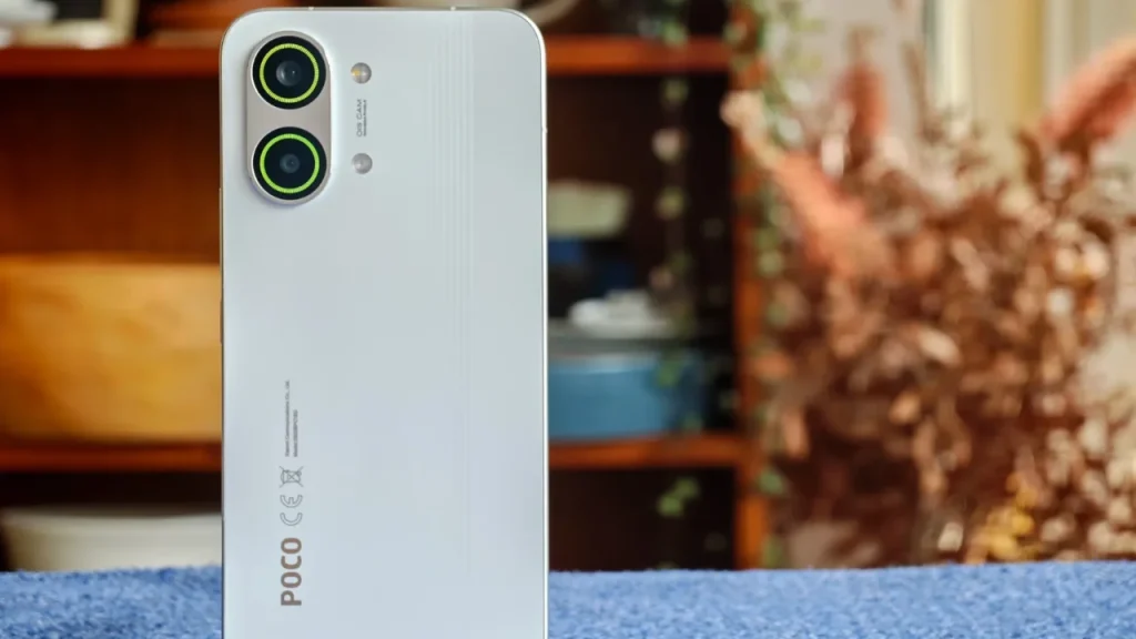 A white Poco X8 Pro smartphone placed on a blue surface with a blurred wooden shelf and flowers in the background.