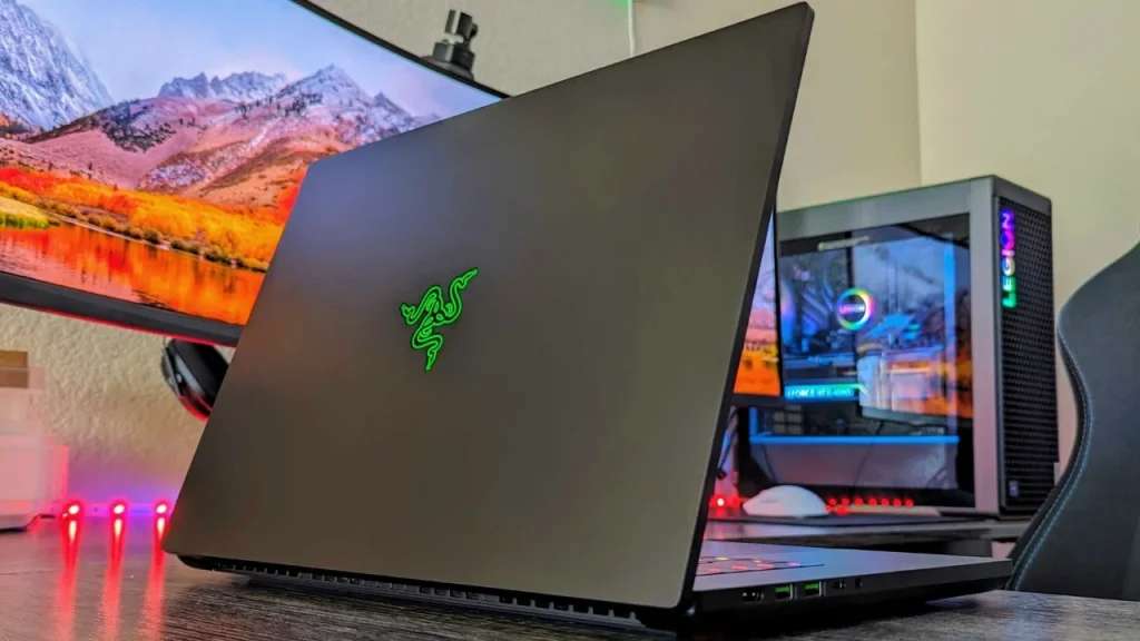 Top 10 Laptop Brands in 2026. A black Razer gaming laptop viewed from the rear, featuring the signature bright green illuminated Razer logo, set up in a gaming environment with RGB lighting.