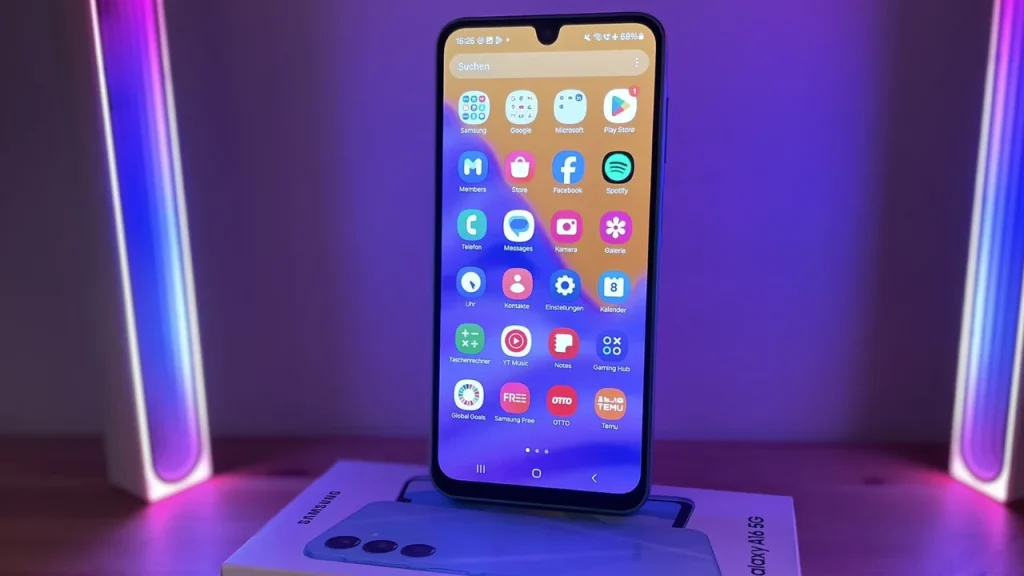 A Samsung Galaxy A16 5G standing upright on its retail box, displaying its home screen with colorful app icons against a purple-lit background.