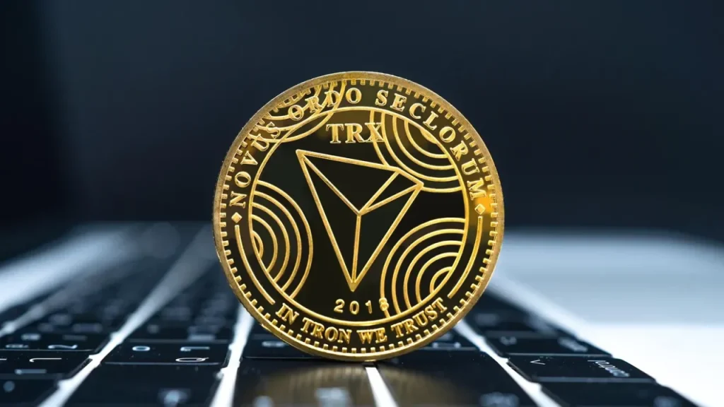 Gold TRON (TRX) coin with a triangular logo and "In Tron We Trust" inscription, resting on a laptop keyboard.